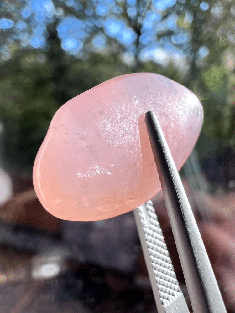 pierre de grenat hydrogrossulaire rose en pierre polie tenu par des brucelles (pinces gemmologiques) sur fond naturel pour illustrer une pierre rose