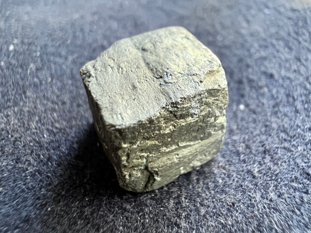 cristal de pyrite naturelle en forme de cube montrant sa couleur metallique doré sur fond de tissu gris pour illustrer la pyrite