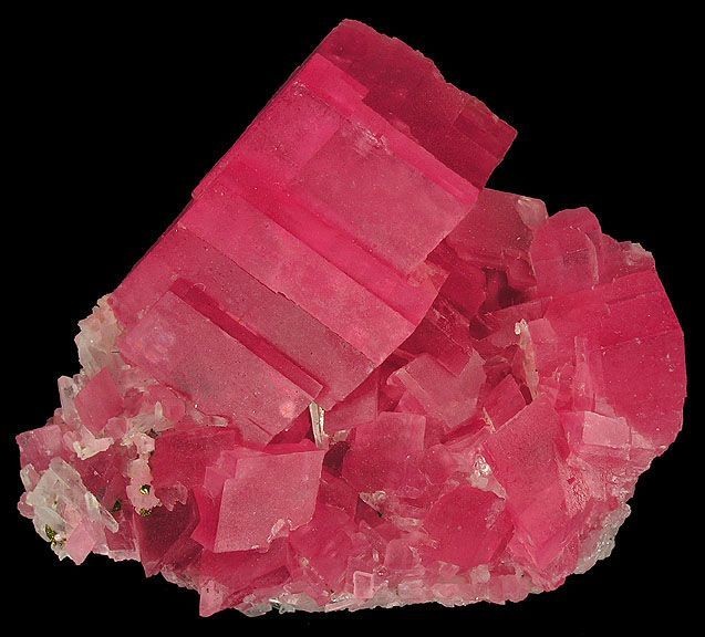 bloc de cristaux de rhodochrosite rosé-rouge montrant des formes ordonnées faites de lignes droites et d'angles qui montrent une structure cristalline. Sa couleur est transparente à translucide et elle est sur un morceau de roche.