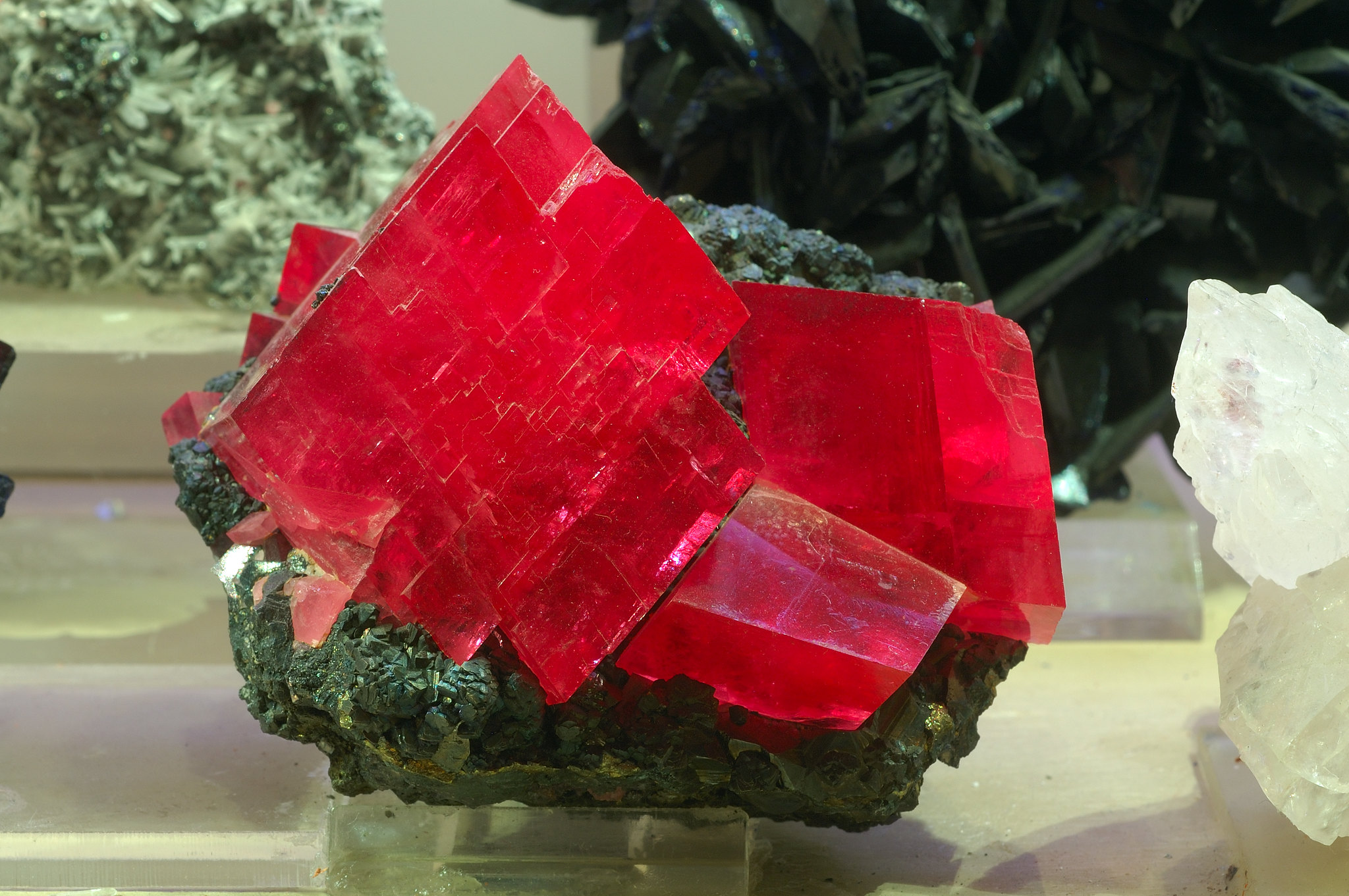 bloc de cristaux de rhodochrosite exposé rosé-rouge montrant des formes ordonnées faites de lignes droites et d'angles qui montrent une structure cristalline. Sa couleur est transparente à translucide et elle est sur un morceau de roche.