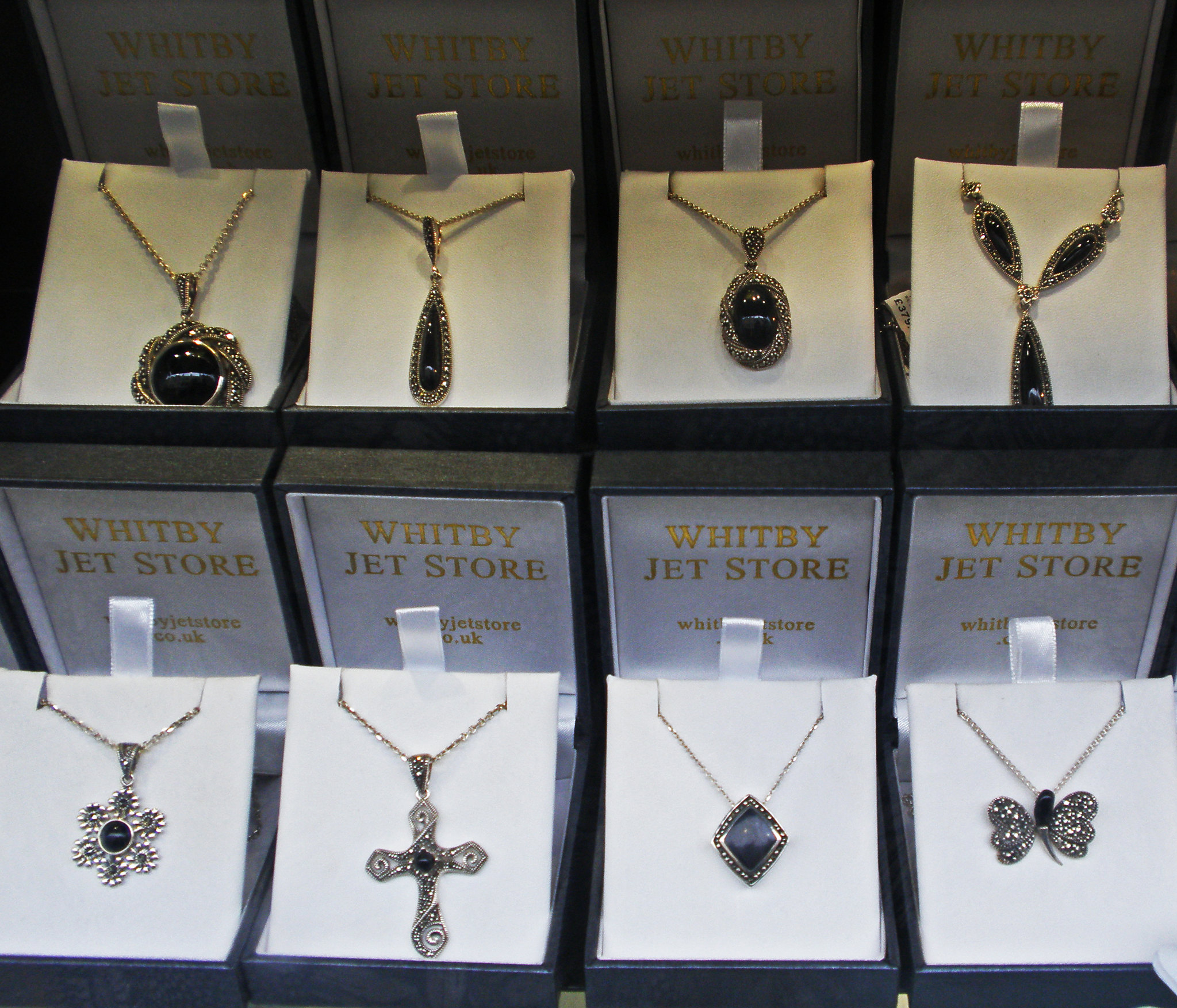 8 boites ouvertes de joiallerie montrant chacune un pendentif en argent en en jais avec des formes de croix, de rond, de losanges et de papillons.