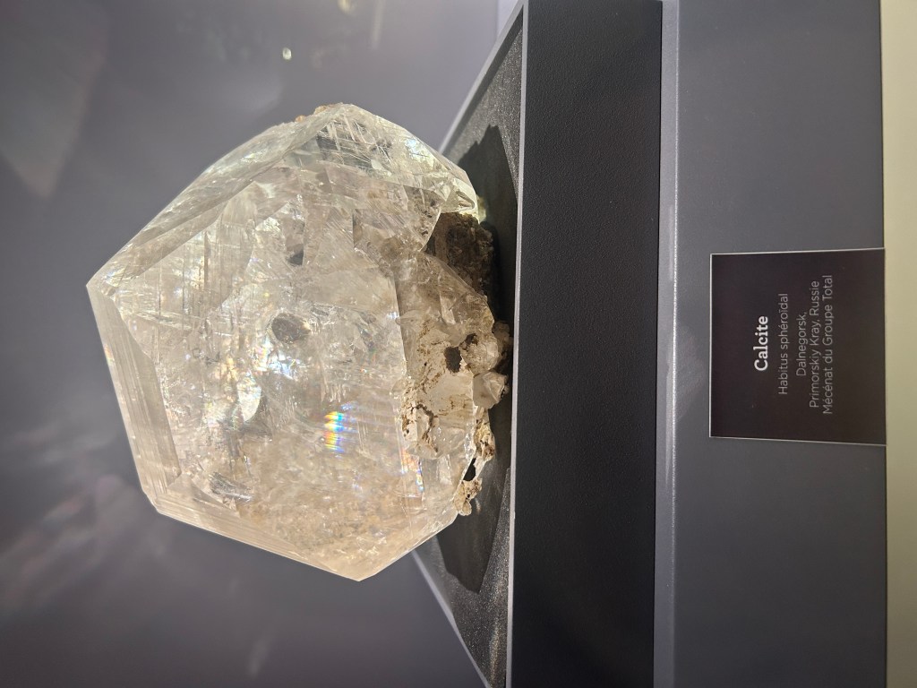 cristal de calcite avec une forme hexagonale fracturée en bas et montrant une couleur blanc à rosé transparent. On vit bien la décomposition de la lumière dans la pierre